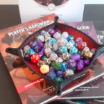 Bronboeken voor Dungeons and Dragons gestapeld onder een dice tray, die op zijn beurt weer gevuld is met allerlei verschillende soorten dobbelstenen.