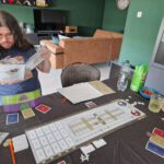 Arran zoekt iets op in het spelregelboek van The King's Dilemma. De tafel staat vol spul voor het spel, drinken en snacks.