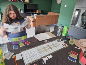 Arran zoekt iets op in het spelregelboek van The King's Dilemma. De tafel staat vol spul voor het spel, drinken en snacks.