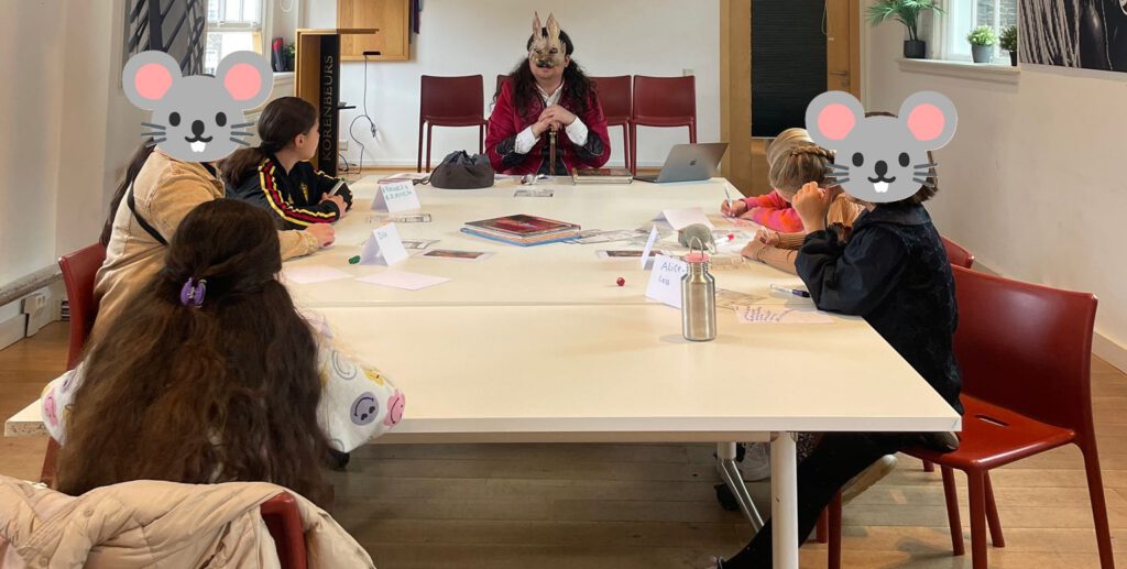 Ik zit verkleed aan het hoofd van de tafel tijdens een sessie Mausritter met kinderen in de bibliotheek van Schiedam